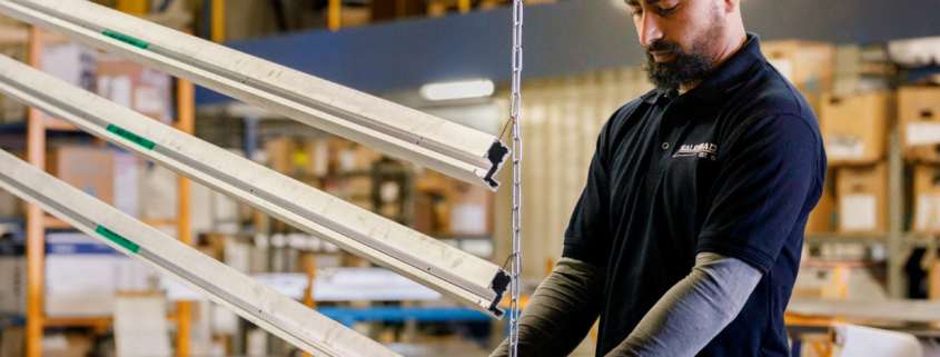 male worker in a dark blue polo shirt and grey undershirt handling long pieces of aluminium in a factory or warehouse setting